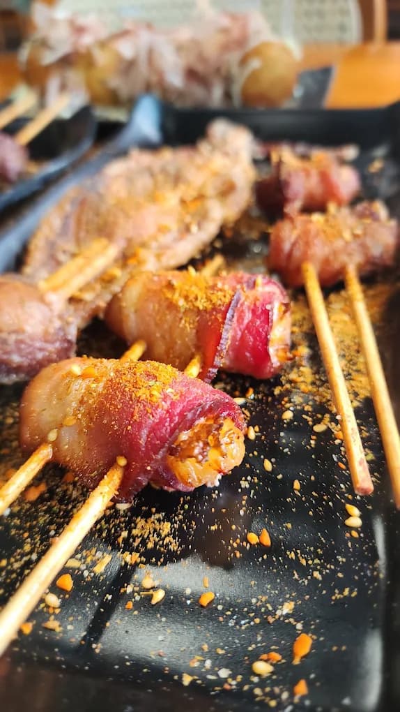 Bacon-wrapped skewers dusted with chili and sesame, close up on a black platter.